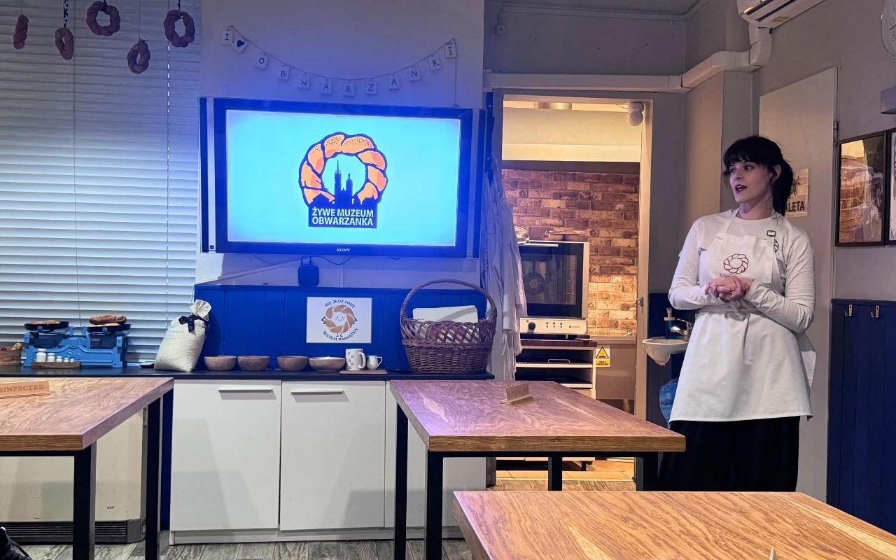 Kobieta w jasnym fartuchu stoi przy ścianie, obok niej znajduje się telewizor z logo Żywe Muzeum Obwarzanka, w pomieszczeniu znajdują się drewniane stoły, naczynia i przybory kuchenne, całość sugeruje prezentację lub warsztaty kulinarne.