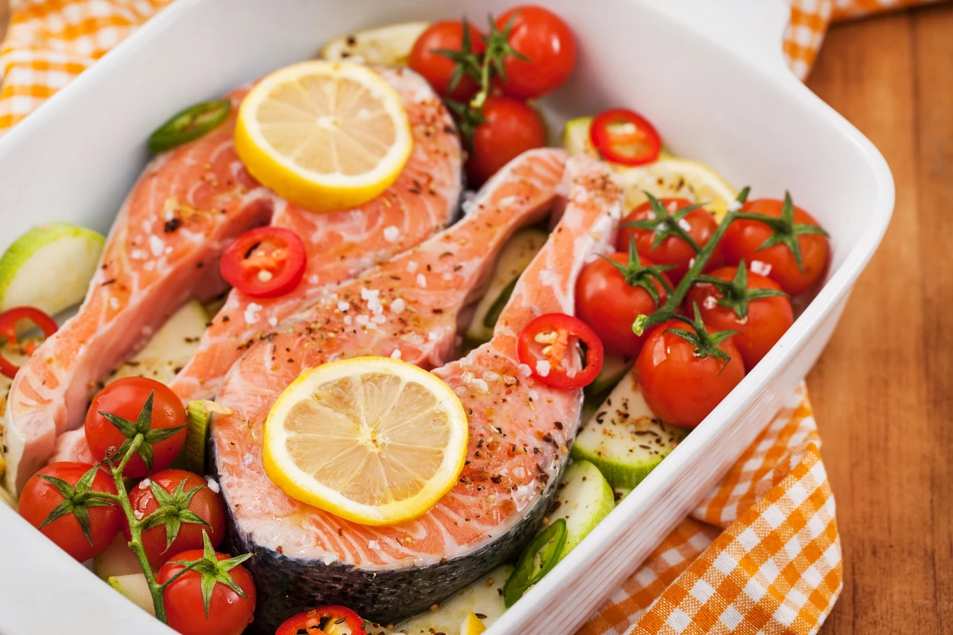 Świeże steki z łososia ułożone w naczyniu do zapiekania, udekorowane plasterkami cytryny, pokrojonymi papryczkami chili, pomidorkami koktajlowymi na gałązce oraz plastry cukinii i przyprawy. Całość przygotowana do pieczenia.