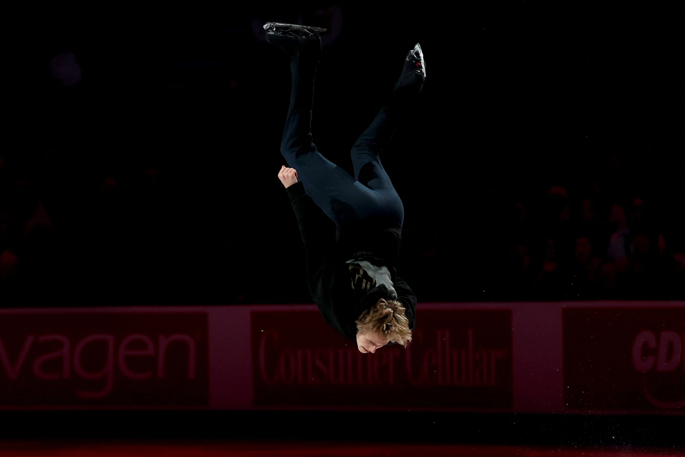 Łyżwiarz figurowy wykonujący efektowny skok z obrotem w powietrzu podczas występu na lodowisku, podkreślony ciemnym tłem i skupionym światłem.