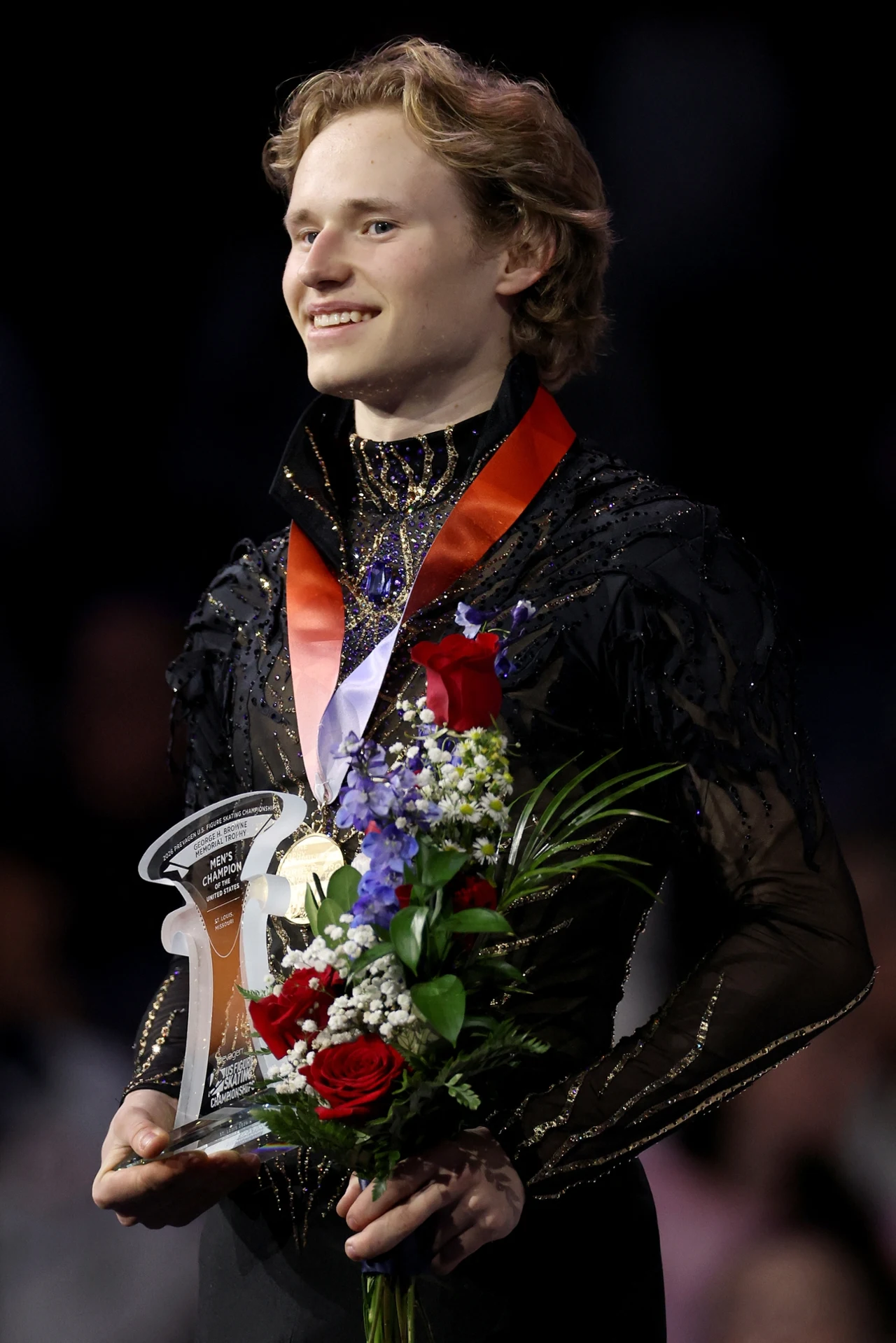 Młody mężczyzna ubrany w elegancki strój sportowy trzyma w rękach bukiet kwiatów i statuetkę, na szyi ma medal, uśmiecha się, tło pozostaje rozmyte i skupia uwagę na nagrodzonym sportowcu.