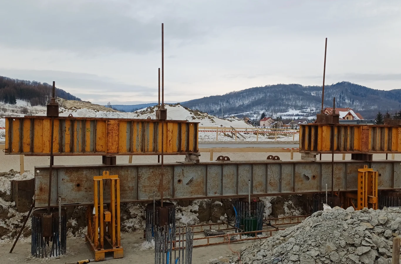 520 ton nacisku. Tak testują fundamenty drogi S19 przy granicy ze Słowacją. Stalowe i betonowe elementy konstrukcyjne mostu w trakcie budowy, żółto-brązowe elementy podpierane stalowymi słupami, w tle ośnieżone wzgórza i kilka domów, zimowy krajobraz.