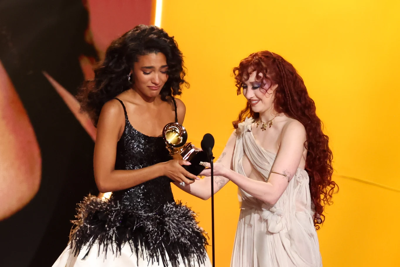 Dwie kobiety stoją na scenie w eleganckich sukniach, jedna wręcza drugiej nagrodę Grammy trzymaną w dłoniach, obie wyrażają silne emocje, tło o ciepłym, żółtym świetle.