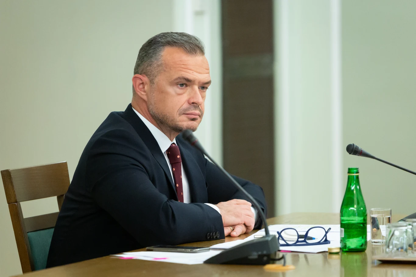 Sławomir Nowak Mężczyzna w garniturze siedzi przy stole konferencyjnym z mikrofonem, zieloną butelką i dokumentami przed sobą, patrząc poważnie w bok.