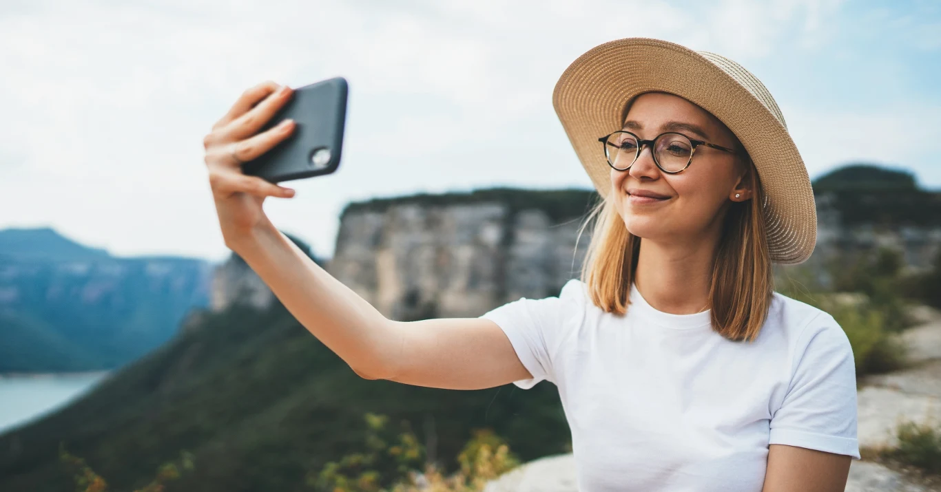 Młoda kobieta w okularach i słomkowym kapeluszu robi selfie na tle wysokich skał oraz zielonego krajobrazu, uśmiechając się do telefonu.
