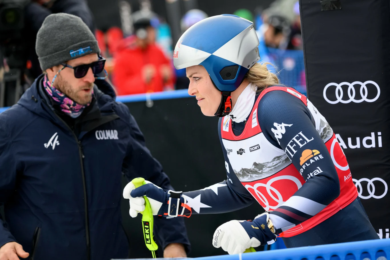 Lindsey Vonn Zawodniczka ubrana w strój narciarski i kask przygotowuje się do startu na trasie zjazdowej, obok stoi trener lub członek ekipy wspierający ją przed rozpoczęciem biegu, w tle widoczne są logo sponsora Audi oraz kibice.