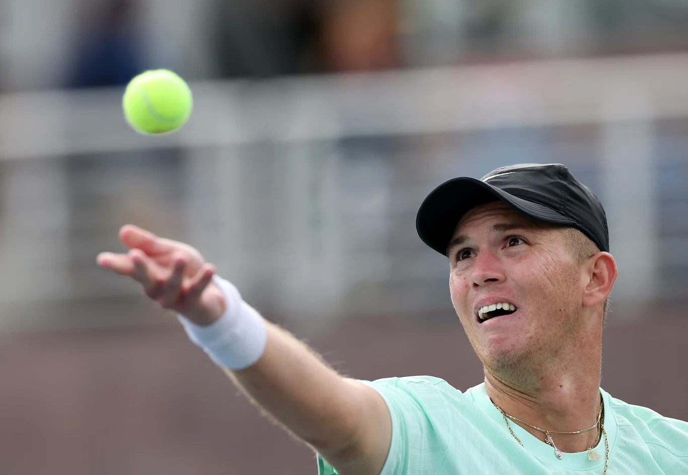 Tenisista w czapce i jasnej koszulce przygotowuje się do serwisu. Na pierwszym planie uniesiona ręka z piłką tenisową, wyraźna koncentracja na twarzy sportowca. W tle rozmazane trybuny.