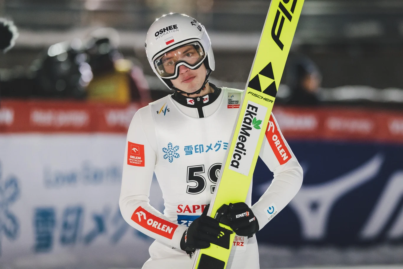 Skoczek narciarski w białym kombinezonie z numerem startowym 59, trzymający żółte narty, stoi na tle banerów sponsorskich na skoczni narciarskiej podczas zawodów.