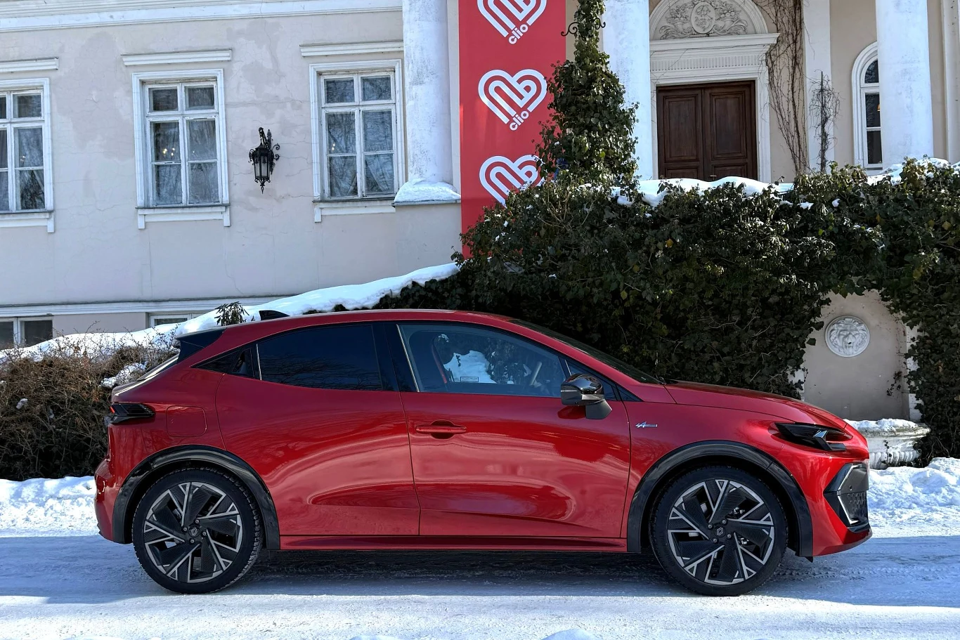 Czerwony samochód elektryczny SUV zaparkowany na tle zabytkowego budynku z klasyczną elewacją, zimowa sceneria, na ścianie widoczne czerwone banery z białym logo i napisem.
