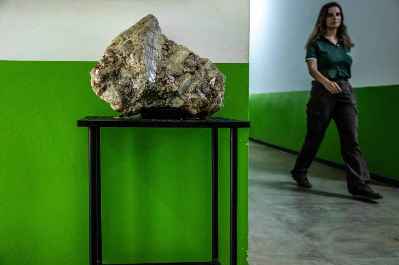 Duży kawałek minerału ustawiony na czarnym metalowym stoliku pod ścianą pomalowaną na biało i zielono, w tle korytarz i przechodząca kobieta w zielonej koszuli oraz ciemnych spodniach.