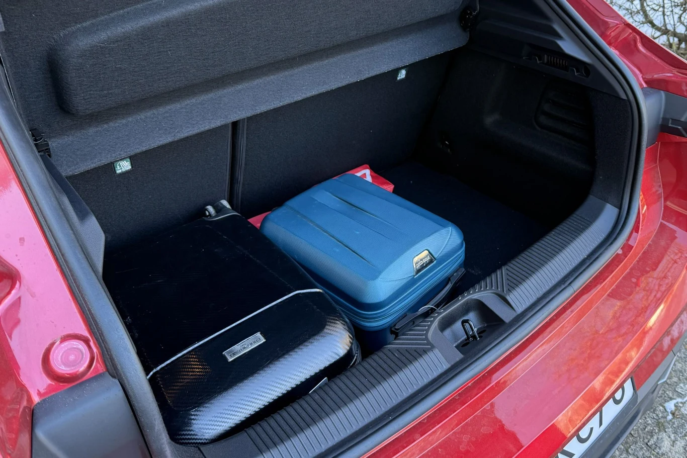 Dwie zamknięte walizki, czarna i niebieska, umieszczone w bagażniku czerwonego samochodu osobowego, widoczne fragmenty wnętrza bagażnika oraz część bagażu podróżnego