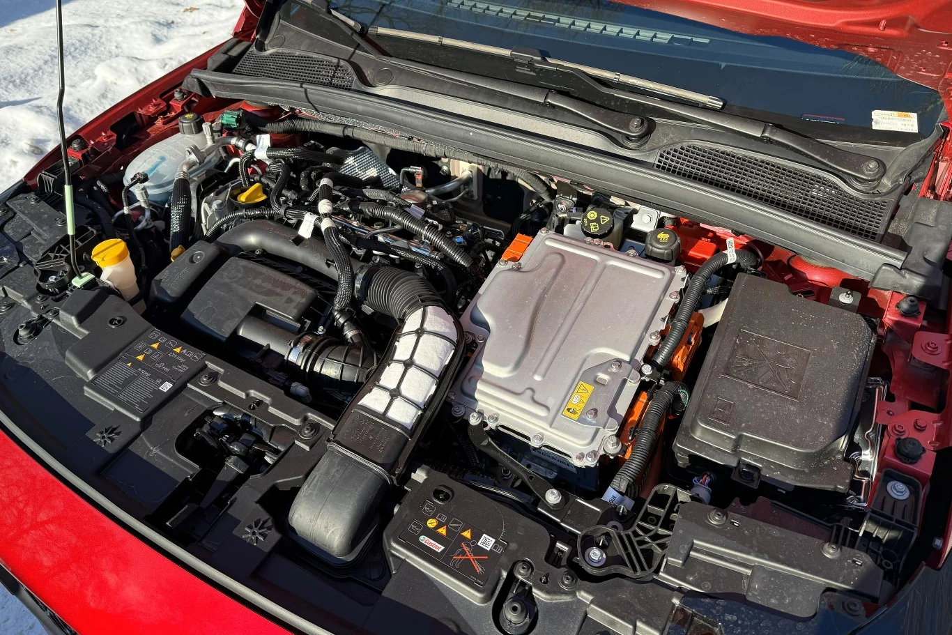 Komora silnika nowoczesnego samochodu osobowego z widocznymi przewodami, modułami elektronicznymi oraz elementami systemu zasilania i chłodzenia. Barwa czerwonego lakieru na krawędzi maski i ślady śniegu w tle.