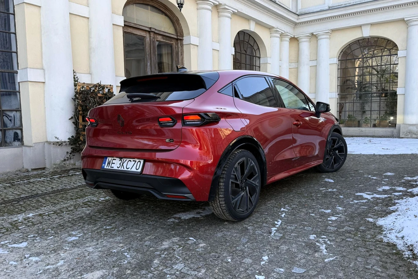 Czerwony samochód osobowy zaparkowany na brukowanej alejce przy historycznym budynku z jasną elewacją i dużymi, łukowatymi oknami.