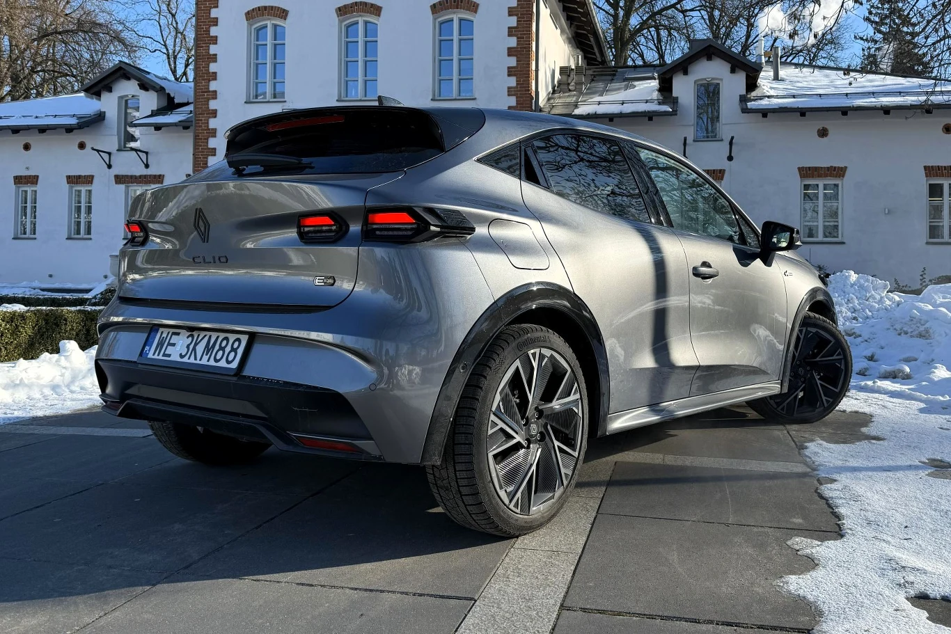 Nowoczesny, srebrny samochód osobowy Renault Clio zaparkowany na podjeździe przed eleganckim, białym budynkiem z czerwonymi detalami. Otoczenie pokryte jest śniegiem, a samochód wyróżnia się wyrazistymi liniami i dużymi felgami.