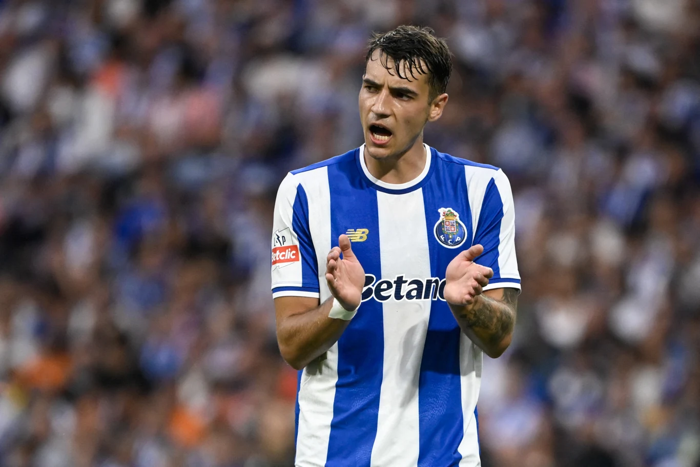 Jakub Kiwior Piłkarz klubu FC Porto w biało-niebieskim stroju sportowym, stojący na boisku, klaszczący w dłonie z widocznym wyrazem koncentracji na twarzy. W tle rozmyty tłum kibiców.