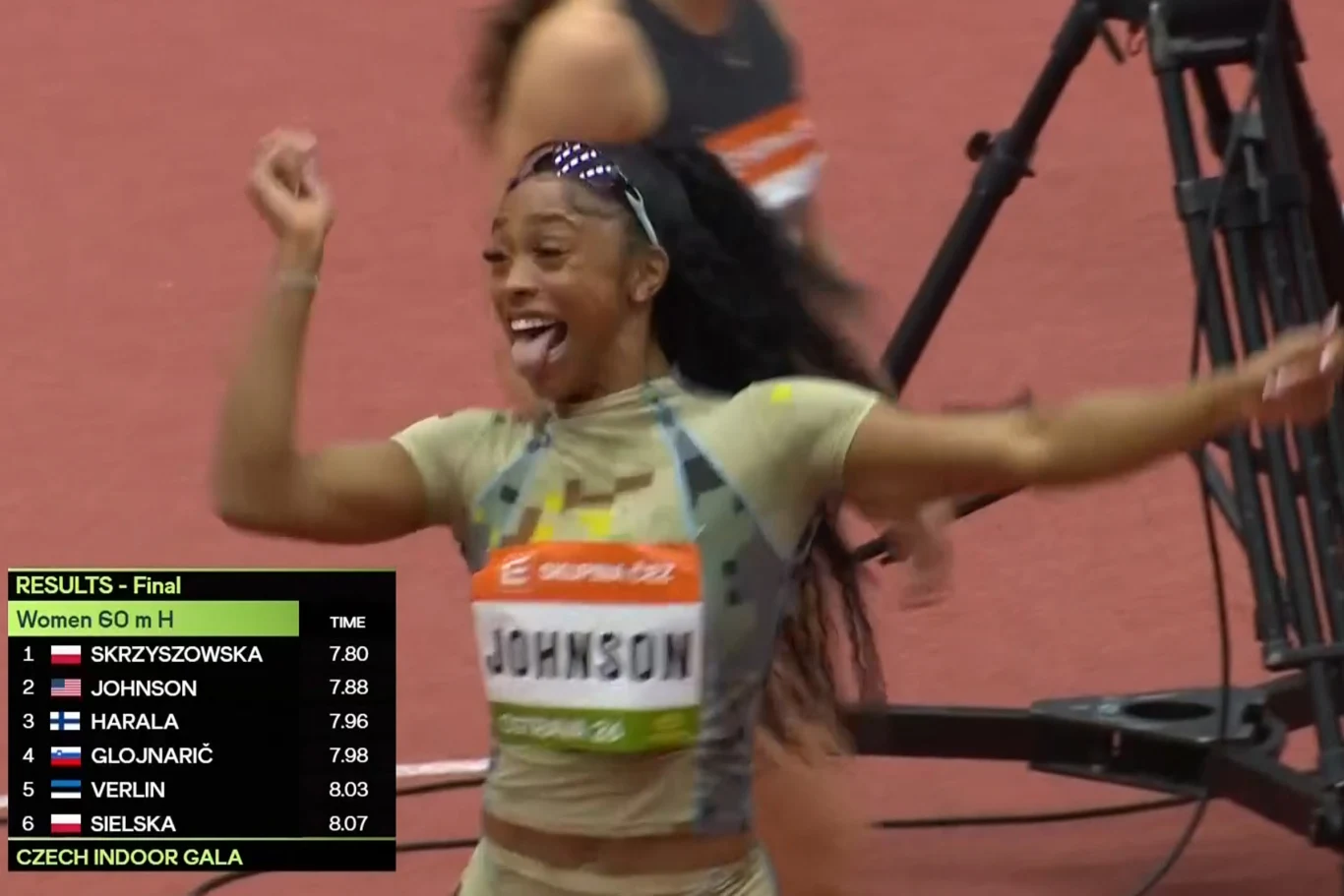 Zawodniczka w sportowym stroju z uśmiechem celebruje wynik na bieżni hali lekkoatletycznej. Na pierwszym planie widoczna jest tablica z wynikami finału biegu na 60 m przez płotki kobiet oraz numer startowy z nazwiskiem 'Johnson'.