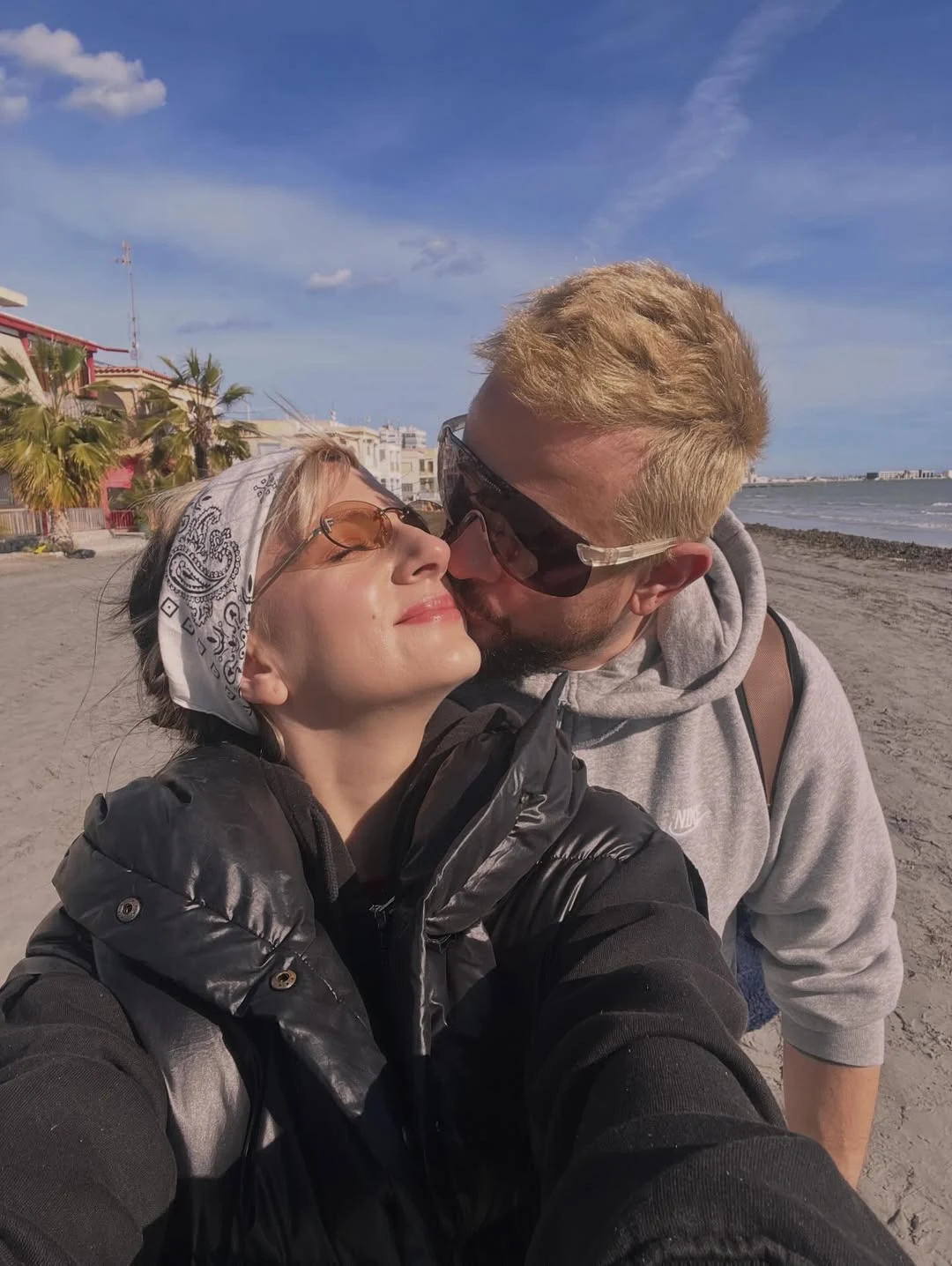 Para obejmująca się na plaży, mężczyzna całuje kobietę w policzek, oboje uśmiechnięci i w okularach przeciwsłonecznych, w tle morze oraz budynki i palmy pod błękitnym niebem.