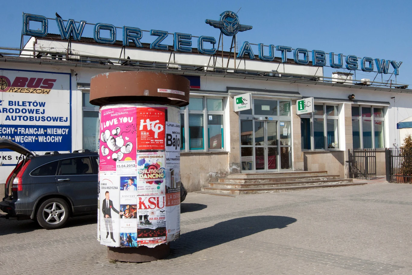 Budynek dworca autobusowego z charakterystycznym napisem na dachu, przed którym stoi słup ogłoszeniowy obklejony kolorowymi plakatami reklamowymi. Z lewej strony widoczne zaparkowane pojazdy, za szybami wnętrze dworca.