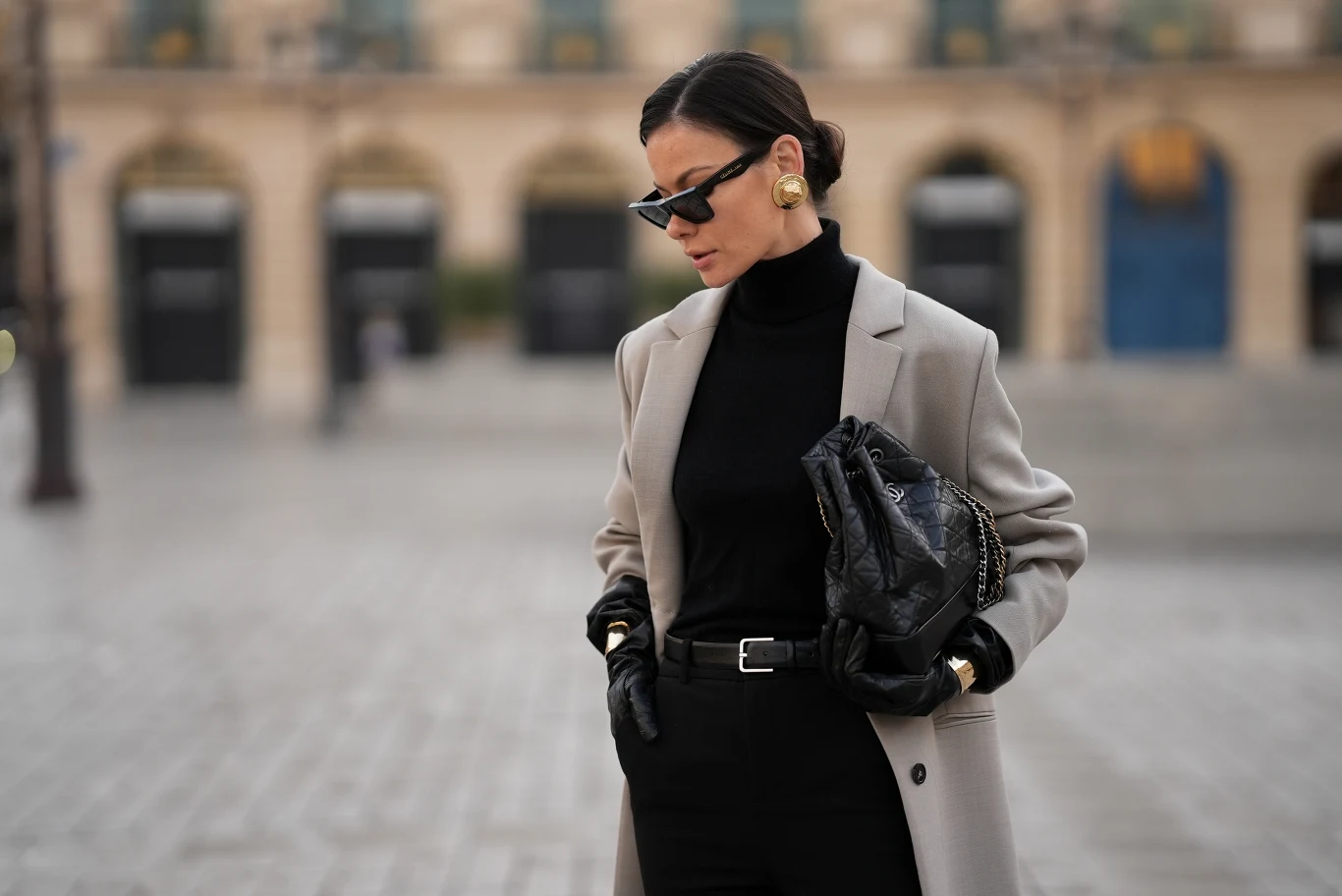 Jak low energy dressing wpływa na nasze samopoczucie? Elegancko ubrana kobieta w czarnym golfie i rękawiczkach, jasnej marynarce oraz okularach przeciwsłonecznych trzyma czarną torebkę na tle miejskiego placu z rozmytym budynkiem w tle.