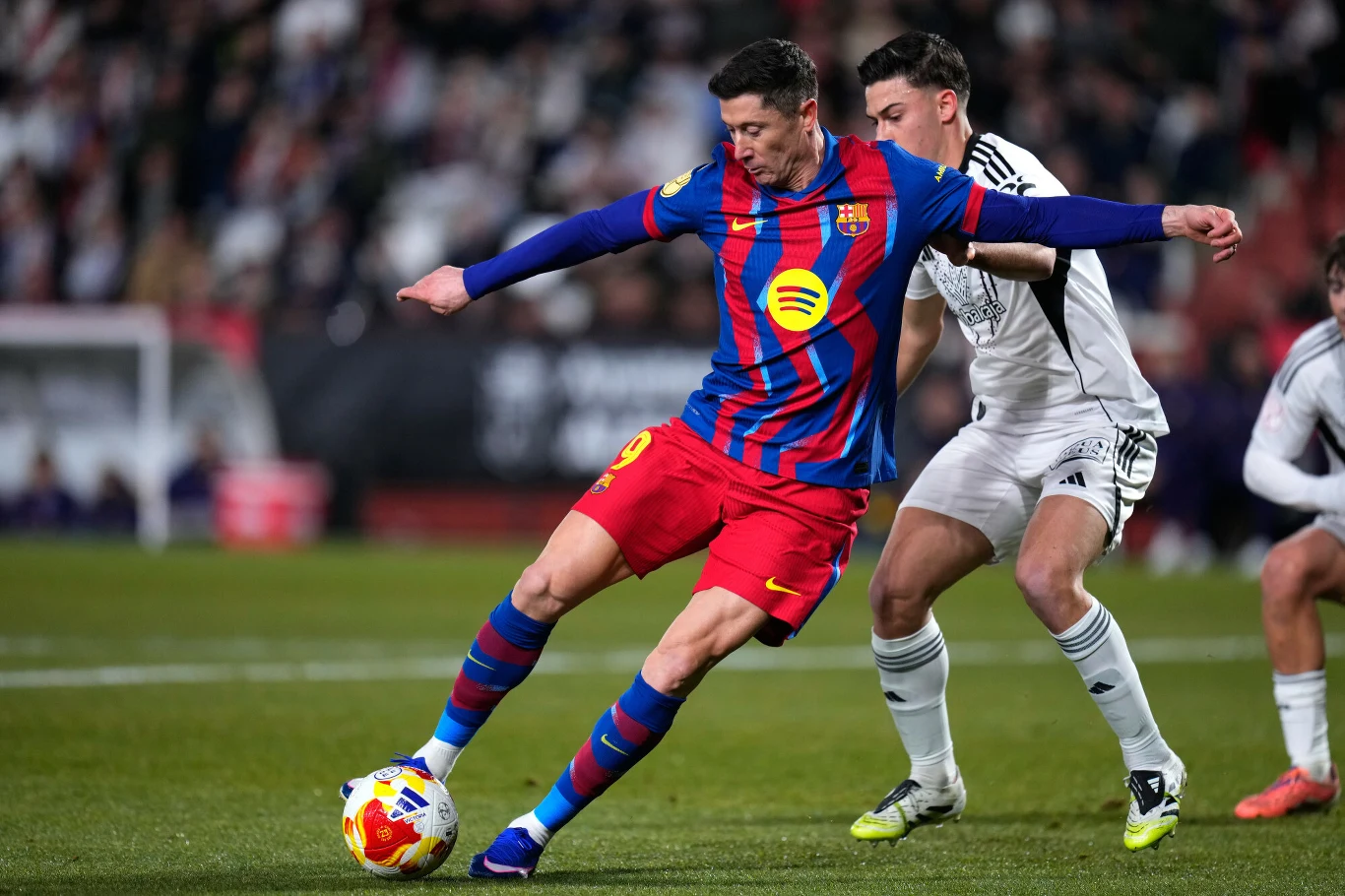 Puchar Króla. Robert Lewandowski podczas meczu z Albacete Balompie Dwóch piłkarzy rywalizuje o piłkę na boisku podczas meczu. Zawodnik w niebiesko-czerwonym stroju kontroluje futbolówkę, podczas gdy przeciwnik w biało-czarnym stroju próbuje go powstrzymać. W tle widoczna rozmazana publiczność na trybunach.