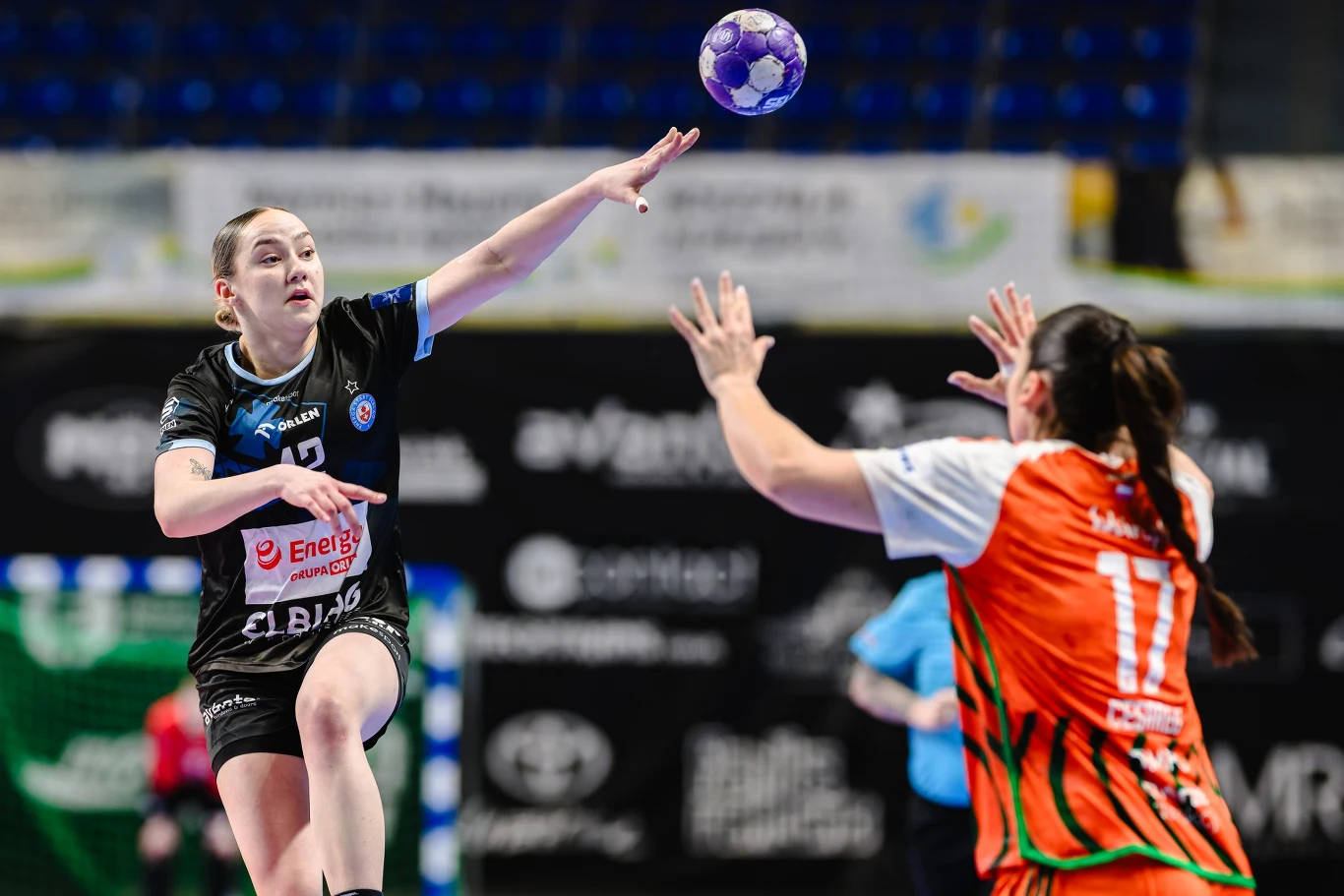 Zawodniczka w czarnym stroju oddaje rzut piłką ręczną w kierunku bramki, próbując ominąć przeciwniczkę w pomarańczowej koszulce, która unosi ręce w geście obronnym. W tle widoczne elementy hali sportowej.