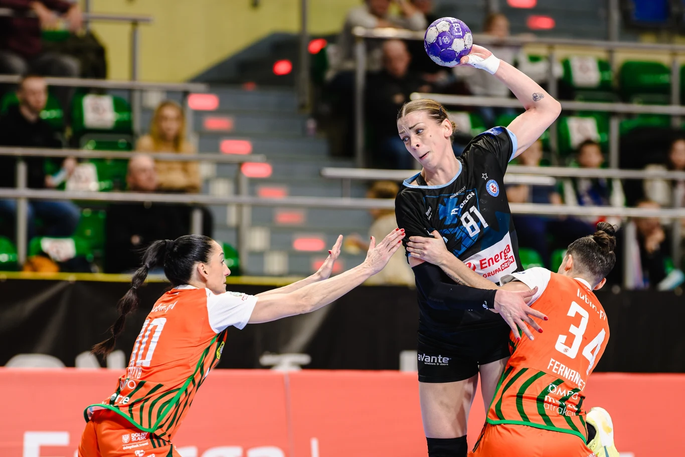 Zawodniczka ubrana w czarny strój z piłką w ręku wykonuje rzut podczas meczu piłki ręcznej, próbując przejść przez dwie rywalki w pomarańczowych strojach na tle trybun z widownią.