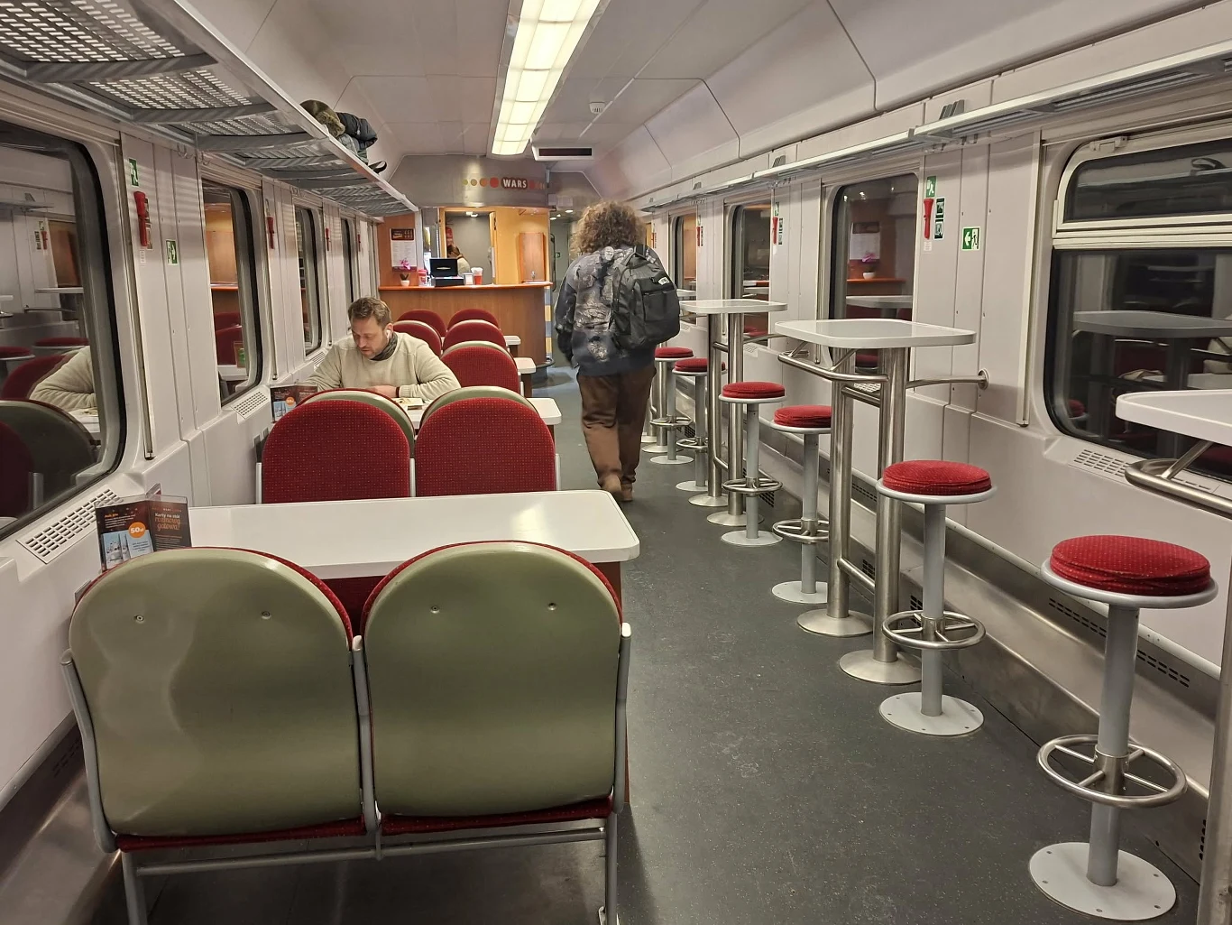 Wnętrze wagonu restauracyjnego w pociągu z czerwonymi i kremowymi siedzeniami oraz stołami; kilku pasażerów zajmuje miejsca, a jeden z nich idzie w stronę wagonu barowego. Jasne oświetlenie i nowoczesny wystrój dominują w scenerii.