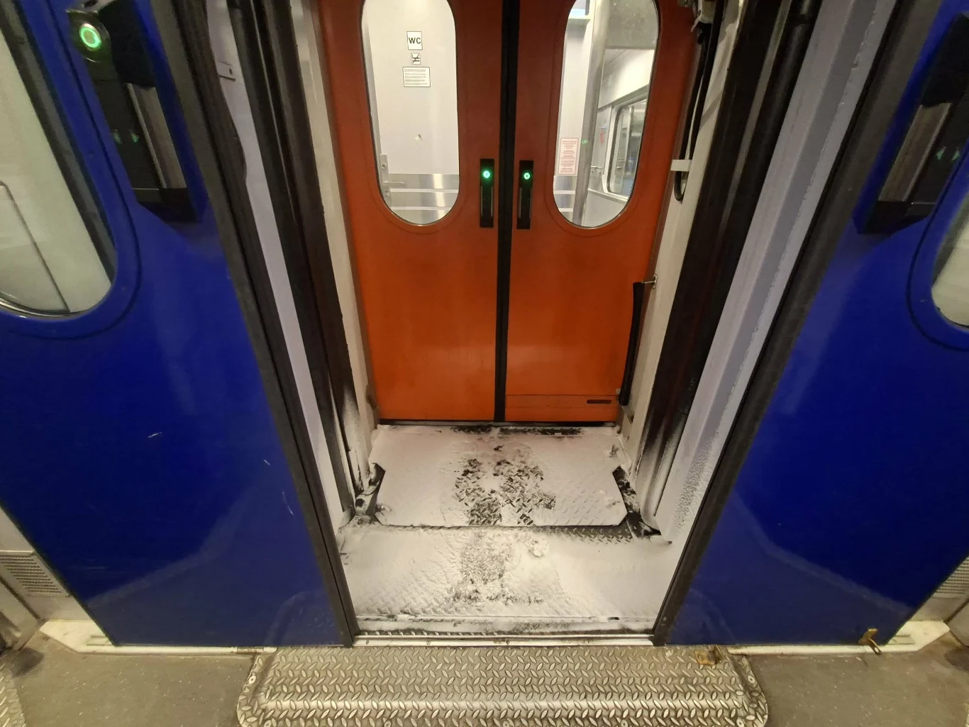 Otwarte drzwi wagonu kolejowego, na progu oraz podłodze widoczna cienka warstwa śniegu, pochodząca zapewne z zewnątrz, z odciśniętymi śladami butów.