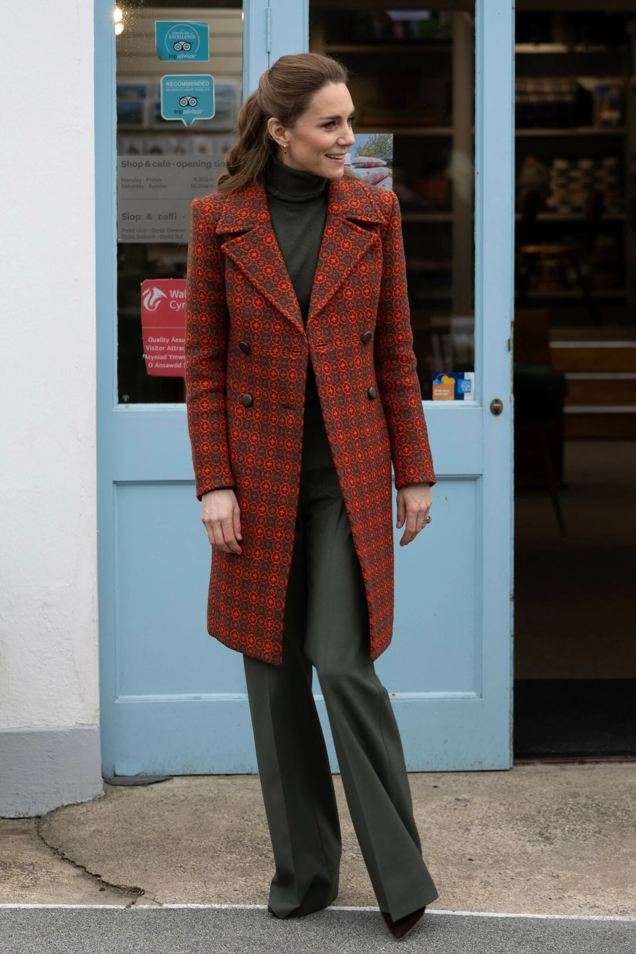 Kobieta ubrana w elegancki, czerwony płaszcz oraz zielone spodnie i golf stoi przed niebieskimi drzwiami wejściowymi do budynku. Ma lekko podniesione włosy, uśmiecha się i patrzy w bok.