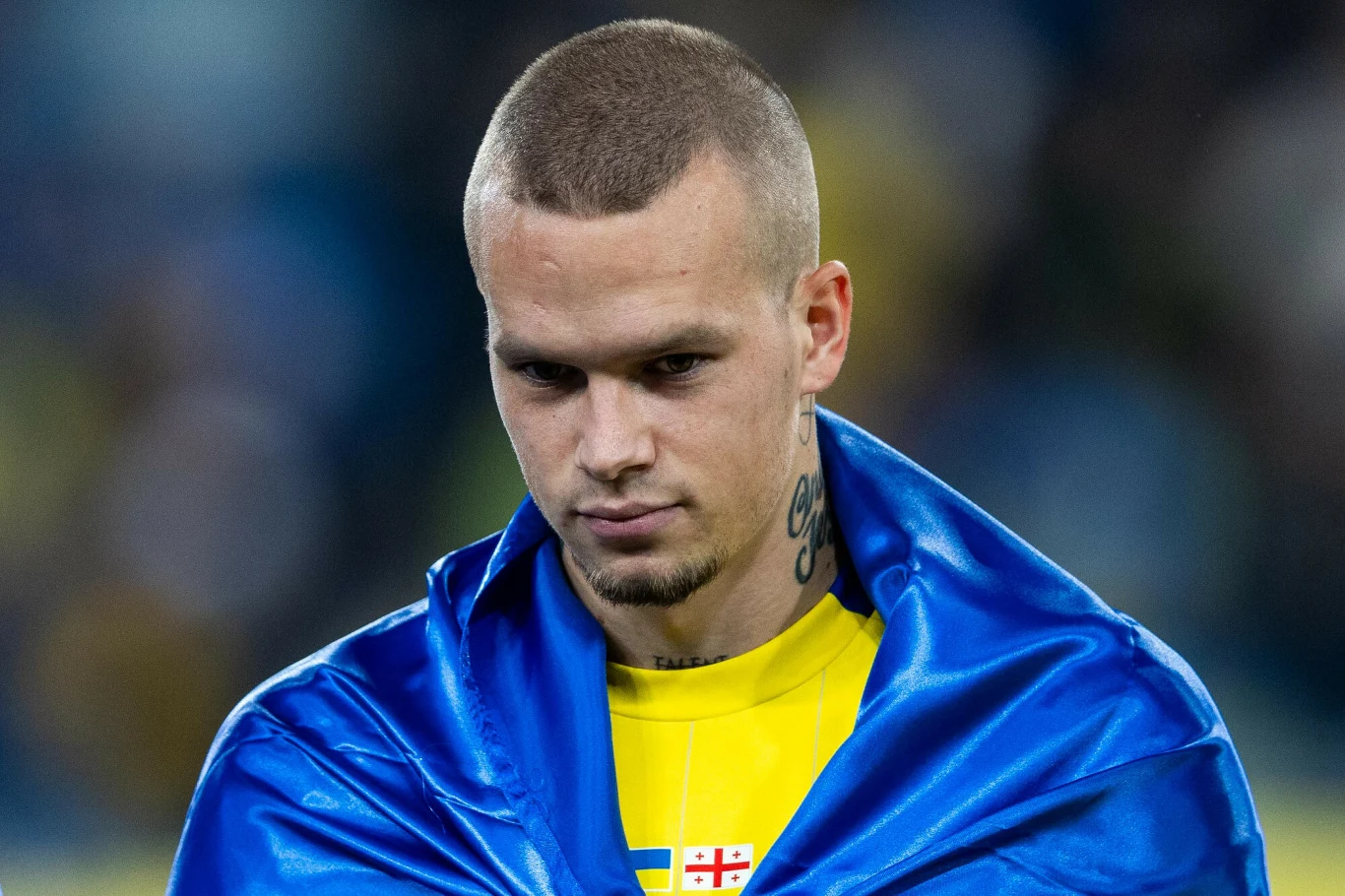 Młody mężczyzna o poważnym wyrazie twarzy, ubrany w żółtą koszulkę z widocznym symbolem narodowym oraz okryty niebiesko-żółtą flagą. Krótkie włosy, tatuaż na szyi, rozmyte tło stadionu.