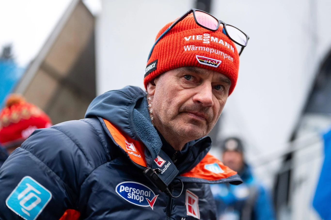 Stefan Horngacher Mężczyzna w czerwonej czapce zimowej z okularami na głowie, ubrany w sportową, granatową kurtkę z licznymi naszywkami reklamowymi, stoi na tle rozmytego zimowego krajobrazu, wśród innych nieostrych postaci.