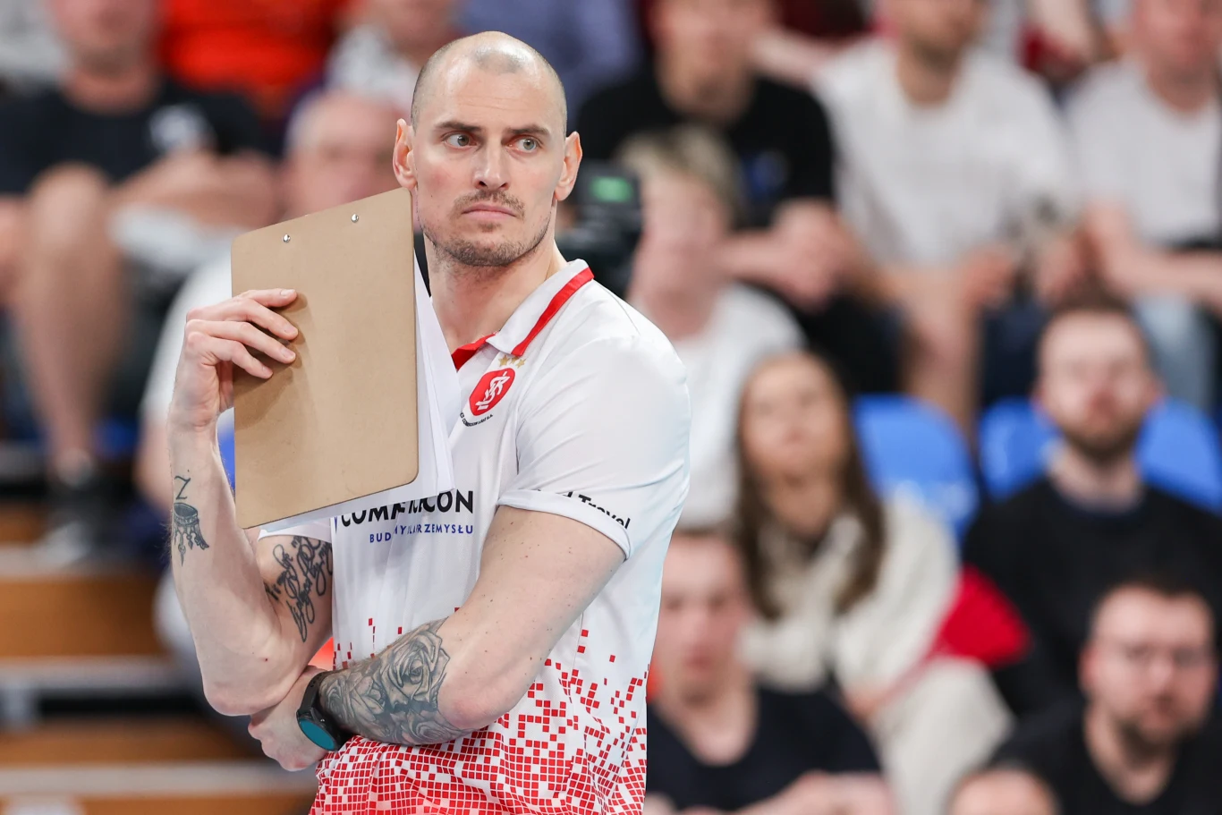 Mężczyzna w sportowej koszulce z logo klubu, trzymający teczkę lub planszę, skoncentrowany podczas wydarzenia sportowego, w tle rozmyte sylwetki widzów na trybunach.