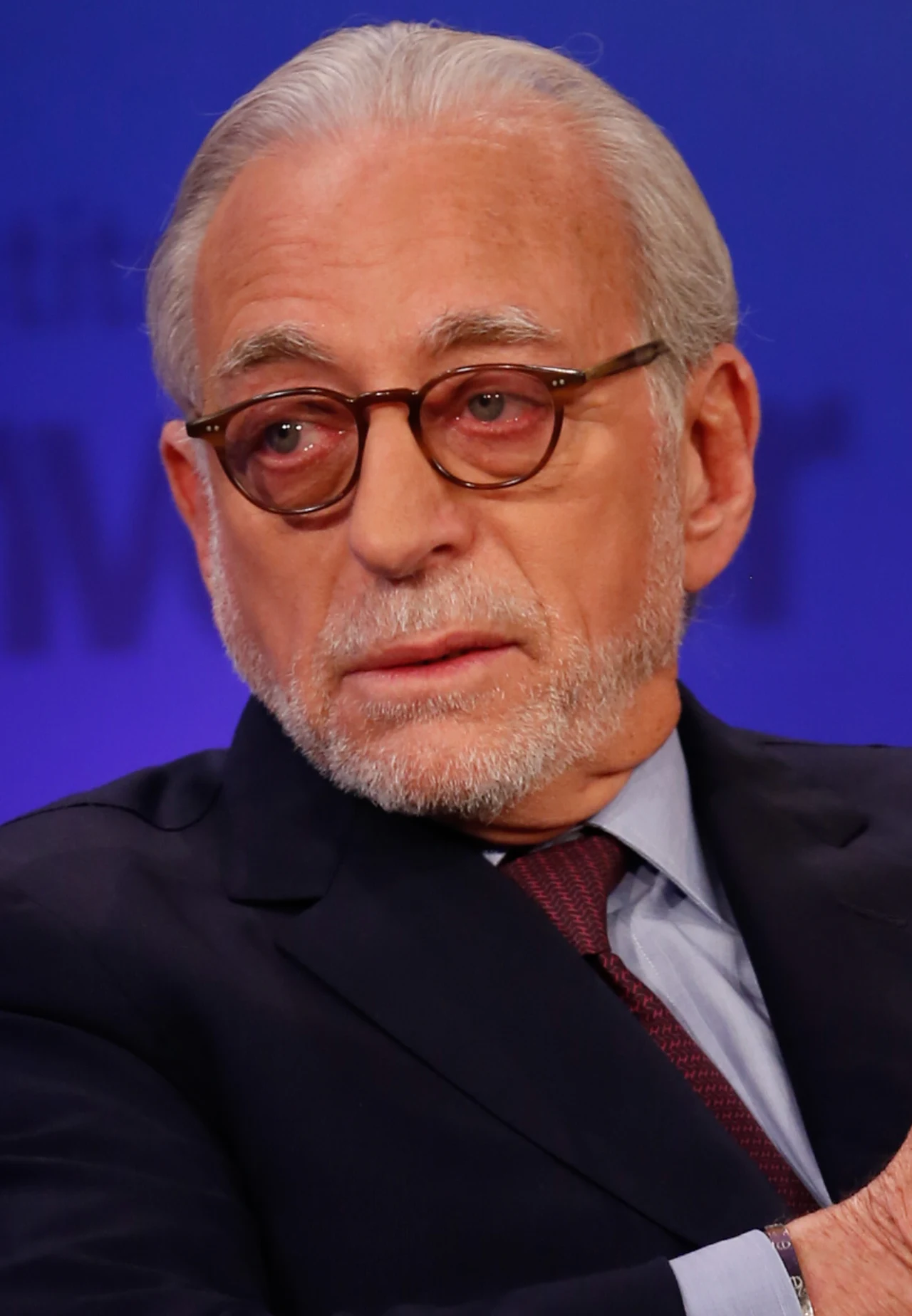 Starszy mężczyzna z siwymi włosami i brodą, w okularach o okrągłych oprawkach, ubrany w elegancki garnitur, krawat i jasnoniebieską koszulę, siedzi na tle jednolitego, niebieskiego tła i patrzy lekko w bok.