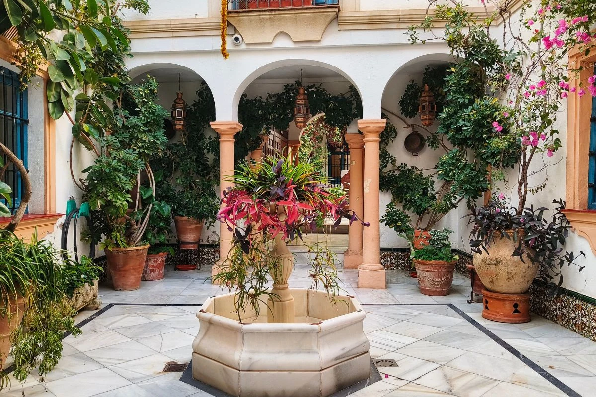 Dziedziniec z marmurową fontanną pośrodku, otoczony donicami z bujną roślinnością oraz trzema łukowymi kolumnami prowadzącymi do zadaszonej części budynku, zabudowania utrzymane w stylu południowoeuropejskim.