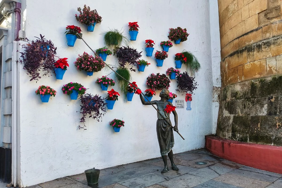Artystyczna rzeźba dziewczynki sięgającej kijem do ściany udekorowanej niebieskimi doniczkami z kolorowymi kwiatami oraz zielonymi roślinami.