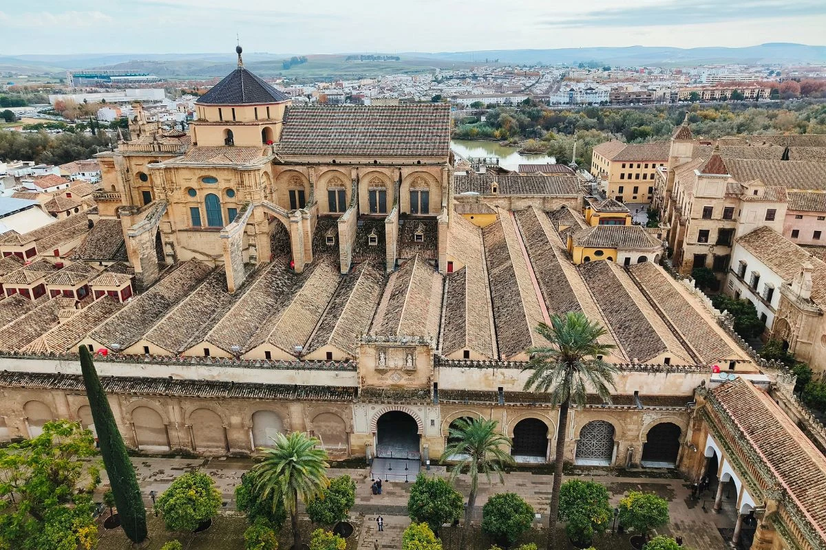 Meczet-katedra w Kordobie widoczny z lotu ptaka, otoczony ogrodem z palmami i cyprysami, z charakterystyczną mieszanką architektury islamskiej i chrześcijańskiej, na tle zabudowy miejskiej i rozległego krajobrazu.