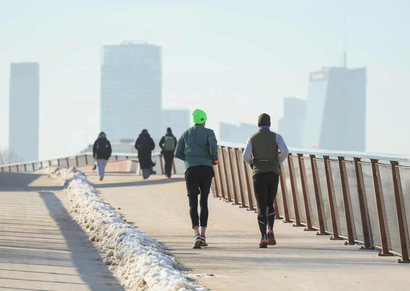 Dwie osoby biegną po szerokiej ścieżce wzdłuż metalowej barierki, przy której leży śnieg. W tle kilku spacerowiczów oraz nowoczesne, wysokie budynki miasta widoczne we mgle.