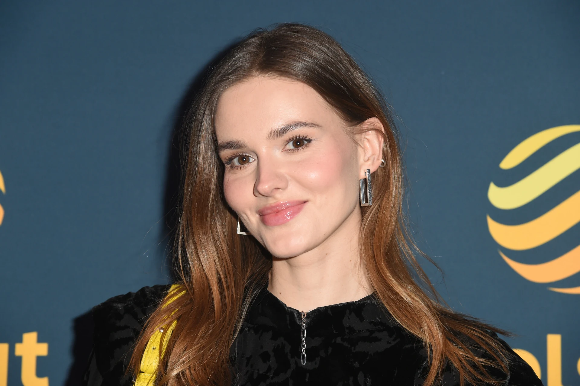 Młoda kobieta z długimi brązowymi włosami uśmiecha się delikatnie do obiektywu. Ubrana jest w czarną, welurową bluzę i nosi geometryczne kolczyki. W tle granatowa ścianka z żółtym logo.