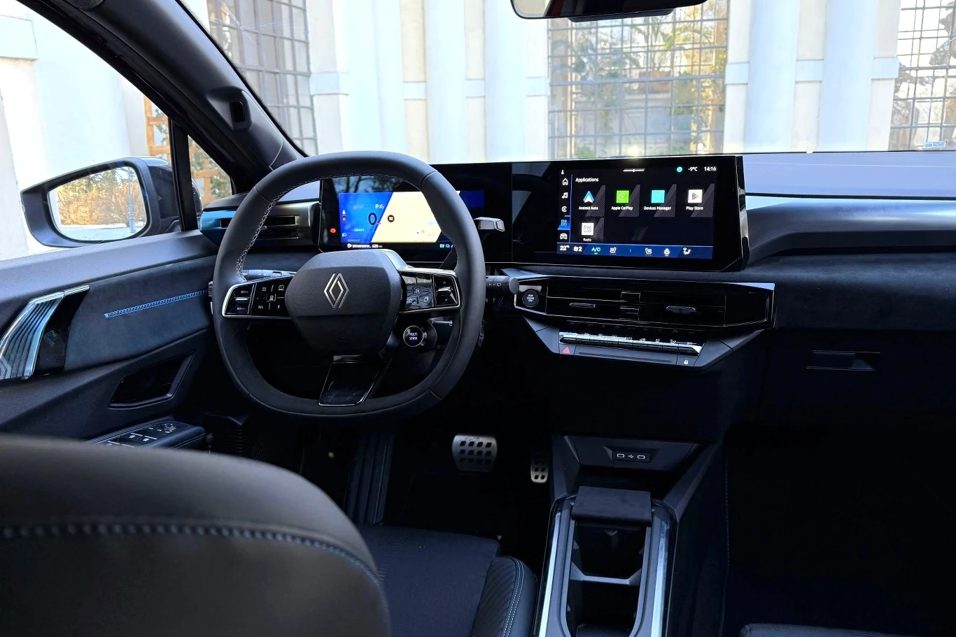 Nowoczesna deska rozdzielcza samochodu z dużym ekranem multimedialnym, cyfrowymi wskaźnikami oraz elegancką kierownicą ze znaczkiem marki Renault. Wnętrze utrzymane w ciemnej kolorystyce, z panoramicznym widokiem na przednią szybę i elementami wykończe...