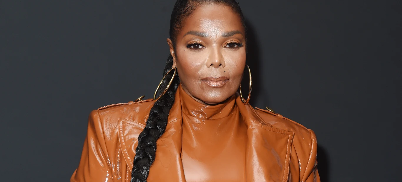Janet Jackson Kobieta w brązowej, skórzanej marynarce i golfie, z długim warkoczem przerzuconym przez ramię, duże złote kolczyki, ciemne tło podkreślające elegancję stylizacji.