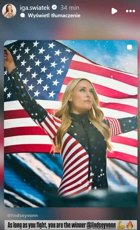 Kobieta ubrana w kostium nawiązujący do flagi Stanów Zjednoczonych stoi z uniesionymi ramionami, w tle widoczna duża flaga amerykańska. Kobieta ma długie blond włosy i patrzy z dumą w dal.