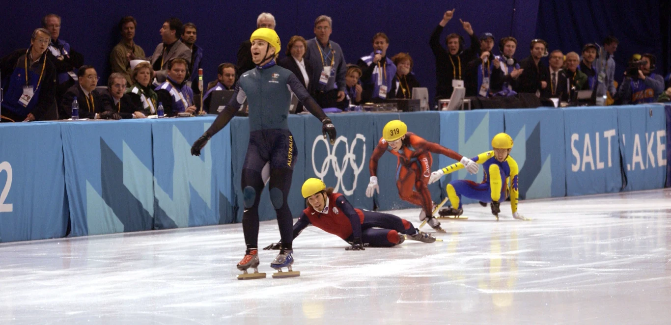 Steven Bradbury w wyjątkowym stylu zdobył pierwszy w historii Australii złoty medal zimowych igrzysk olimpijskich Zawodnik jazdy szybkiej na łyżwach unosi ręce w geście triumfu, za jego plecami inni uczestnicy wyścigu upadają na lód. W tle widoczni kibice oraz sędziowie przy bandzie lodowiska. Widoczne logotypy olimpijskie i napis Salt Lake.