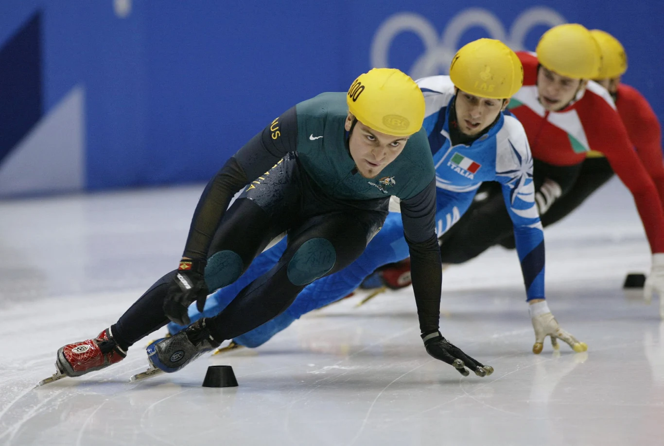 Steven Bradbury na zimowych igrzyskach olimpijskich 2002 w Salt Lake City Łyżwiarze szybcy w kolorowych kombinezonach i żółtych kaskach walczą o prowadzenie na lodowej tafli podczas zawodów. Na pierwszym planie zawodnik skupiony, pochylony w dynamicznym zakręcie, tuż za nim trzech innych uczestników wyścigu.