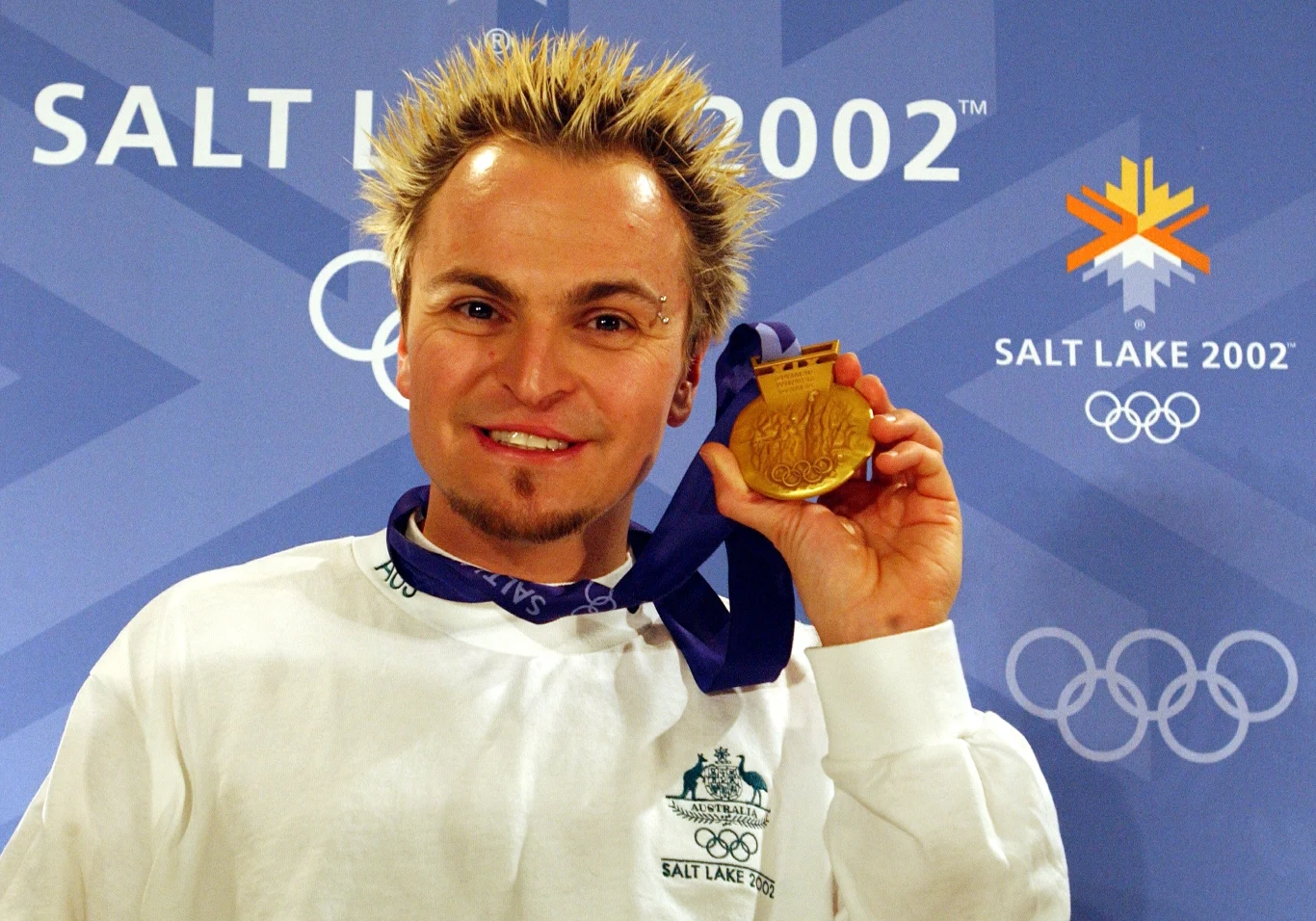 Steven Bradbury ze złotem olimpijskim Sportowiec z blond włosami o kolczystej fryzurze uśmiecha się i trzyma złoty medal olimpijski na tle logo Zimowych Igrzysk Olimpijskich Salt Lake City 2002.