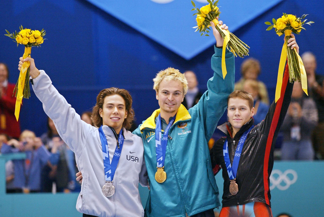 Steven Bradbury ze złotem igrzysk olimpijskich. Obok niego na podium stanęli Apolo Anton Ohno (srebro) oraz Mathieu Turcotte (brąz) Trzech sportowców z medalami olimpijskimi na szyjach, stojących na podium i trzymających bukiety żółtych kwiatów, wyraźnie cieszących się ze swoich osiągnięć.