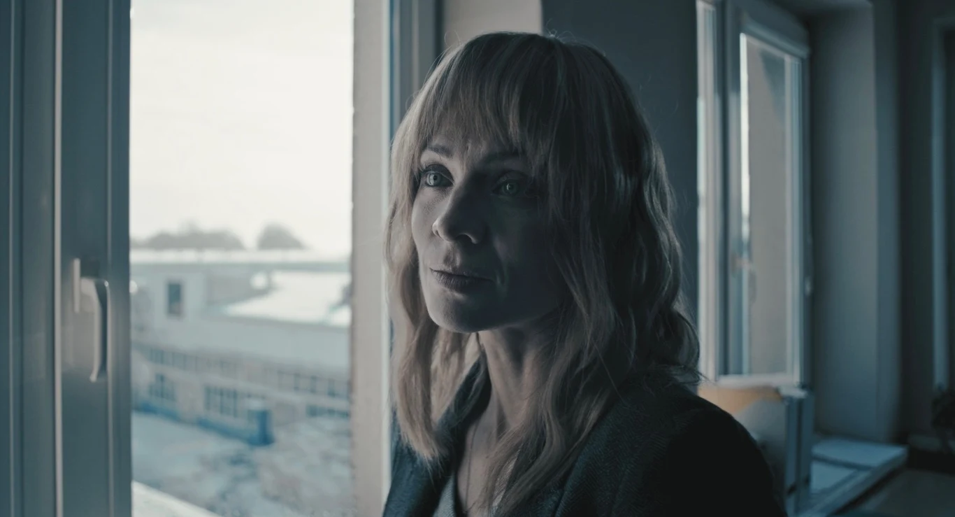 Kobieta o jasnych włosach patrzy poważnie przez okno, w tle widoczne zabudowania i szare światło dzienne tworzące melancholijną atmosferę.