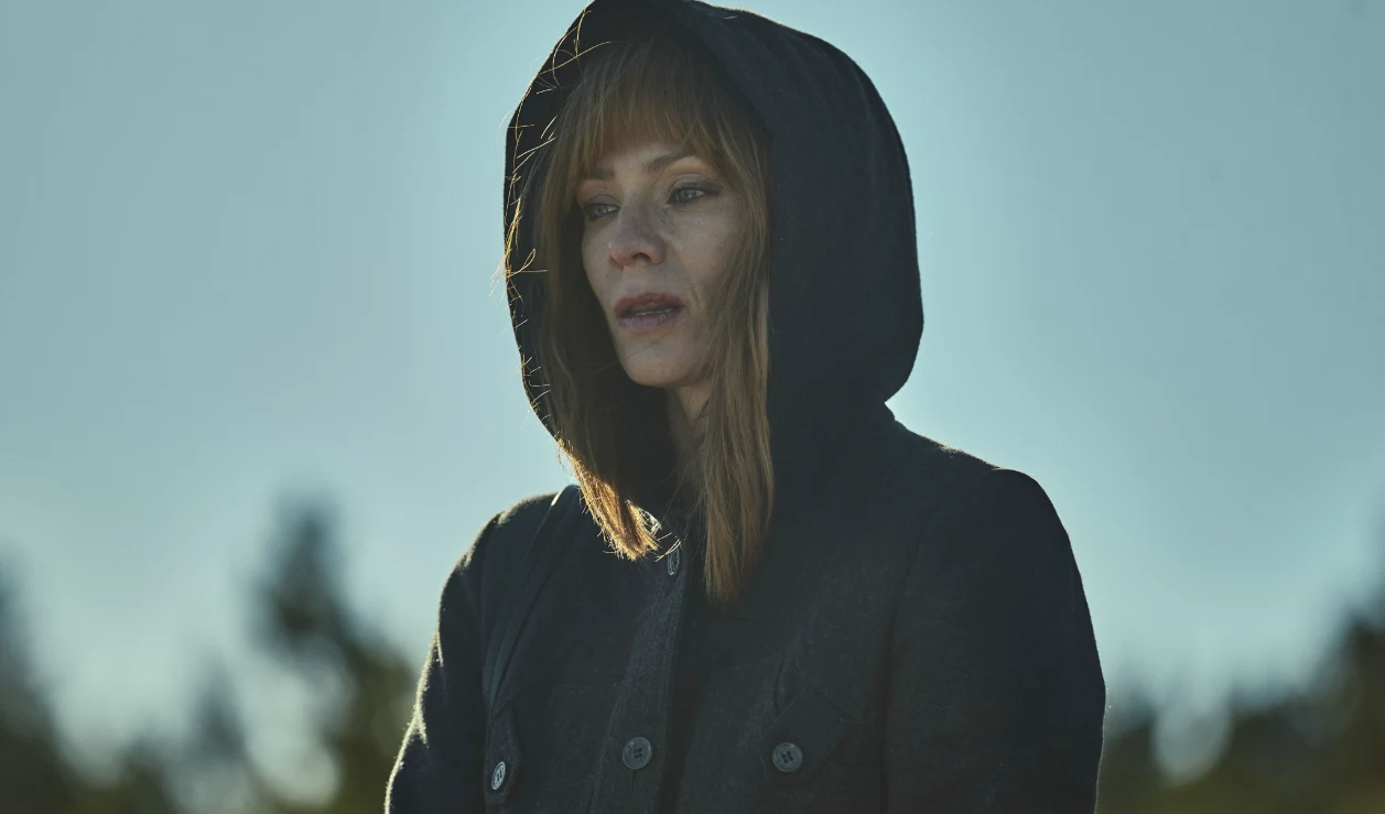 Kobieta z rudymi włosami ubrana w czarną kurtkę z kapturem stoi na zewnątrz. Jej wyraz twarzy wyraża zamyślenie lub smutek, w tle rozmyte, jasne niebo i zarys drzew.
