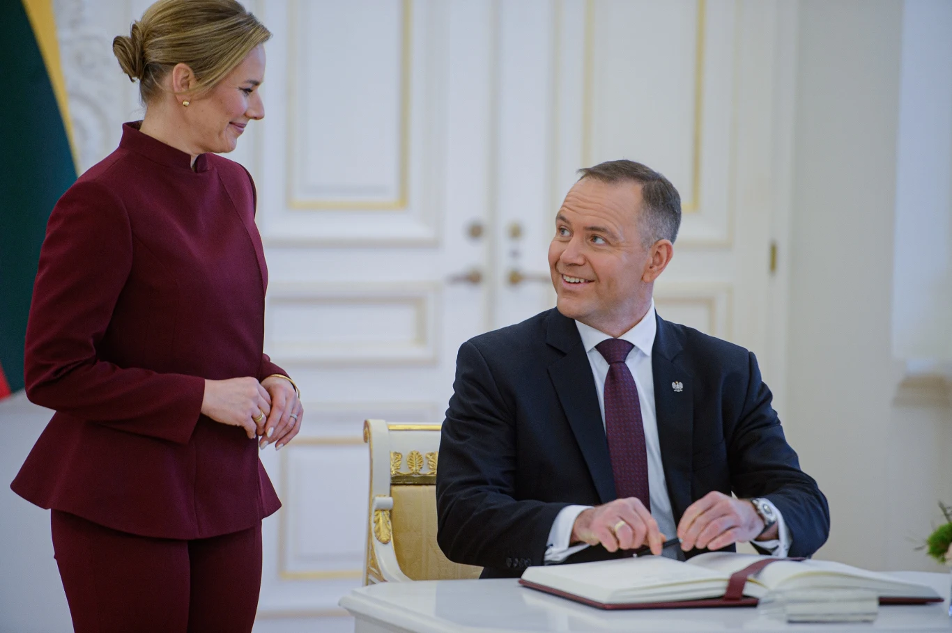 Para prezydencka przyjęła w Pałacu Prezydenckim Dorotę Rabczewską Dwóch elegancko ubranych ludzi w oficjalnym wnętrzu, jeden mężczyzna siedzi przy stole i podpisuje dokument, uśmiechając się do stojącej kobiety.