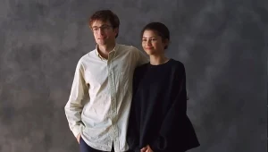 Młody mężczyzna w białej koszuli i okularach oraz młoda kobieta w ciemnym swetrze stoją razem na tle szarej, jednolitej ściany, uśmiechnięci, pozujący do zdjęcia.