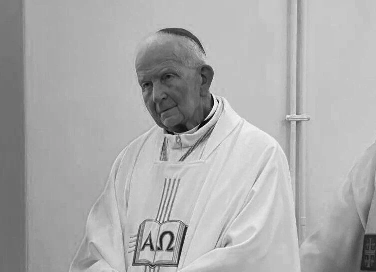 Nie żyje bp Julian Wojtkowski Starszy duchowny w białej szacie liturgicznej stoi na tle jasnej ściany, z wyrazem zamyślenia na twarzy. Na jego ornacie widnieje symbol alfa i omega.