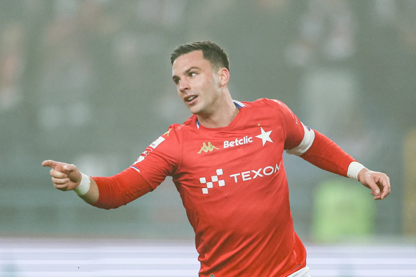 Piłkarz w czerwonym stroju sportowym z logo sponsorów wykonuje gest wskazania ręką podczas meczu, na rozmytym tle widać stadion.